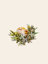 The Peony Birth Flower Ring by CAMELLIA showcases a carved figure surrounded by green and white preserved flowers on a cream background, handcrafted in New York.